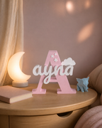 Lettre décorative rose avec le prénom Ayna intégré, posée sur une table de chevet dans une chambre d’enfant chaleureuse.